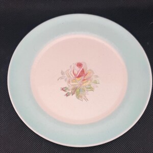 May include: A round, vintage-style plate with a light blue rim and a pink center. The center features a detailed floral design of a rose with red, yellow, and green hues. The plate is likely made of ceramic or porcelain.