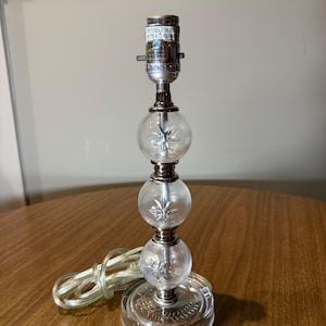 May include: A clear glass table lamp with a decorative design. The lamp features three spherical glass sections with a starburst pattern, connected by metal accents. The base is round with a leaf pattern. A white cord extends from the base.