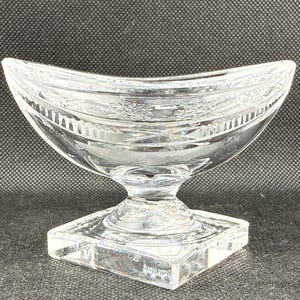 May include: A clear, decorative glass bowl with a curved, boat-like shape. The bowl sits on a square base and has a detailed floral pattern around the rim. The bowl is made of clear glass.