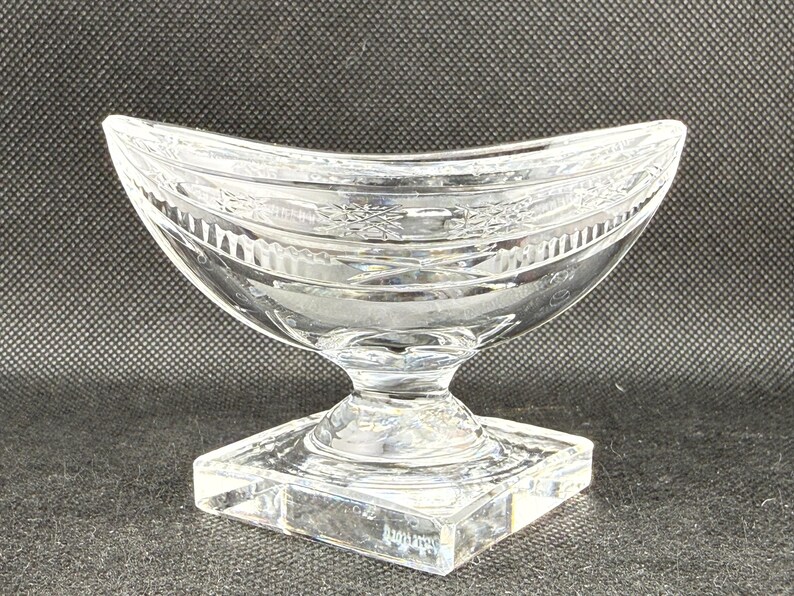 May include: A clear, decorative glass bowl with a curved, boat-like shape. The bowl sits on a square base and has a detailed floral pattern around the rim. The bowl is made of clear glass.