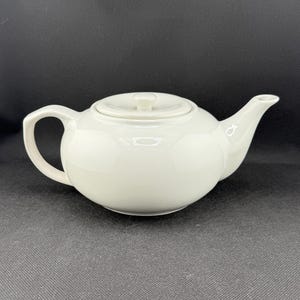 May include: A white ceramic teapot with a lid. The teapot has a round body and a spout on the right side.