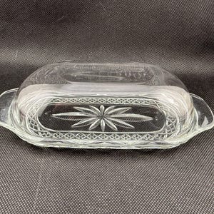 May include: Clear glass butter dish with a domed lid and a rectangular base. The base features a starburst design in the center and decorative etched patterns. The dish has handles on each end.