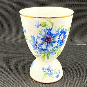 May include: White ceramic egg cup with a gold rim and a floral design featuring blue and purple cornflowers. The egg cup has a flared top and a pedestal base, creating a classic, elegant look.