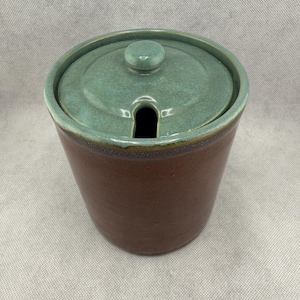 May include: A brown ceramic jar with a teal lid. The jar has a cylindrical shape and a small opening in the lid for a spoon. The jar is likely used for storing condiments or other food items.