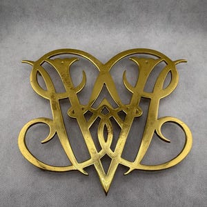 May include: A gold-colored metal decorative monogram featuring the intertwined letters "AW" in an ornate, stylized design. The monogram has a heart-like shape with curved flourishes and a pointed bottom. The metal has some signs of age and wear.