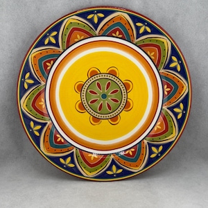 May include: A decorative ceramic plate with a vibrant floral design. The plate features a yellow centre with a detailed floral pattern, surrounded by concentric rings of white, orange, and blue. The outer rim has a repeating pattern of colourful shapes.