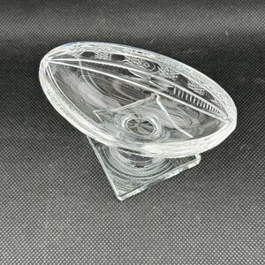 May include: A clear, oval-shaped glass dish with a decorative etched pattern around the rim. The dish sits atop a triangular base, creating a pedestal design. The glass has a reflective quality, catching light.
