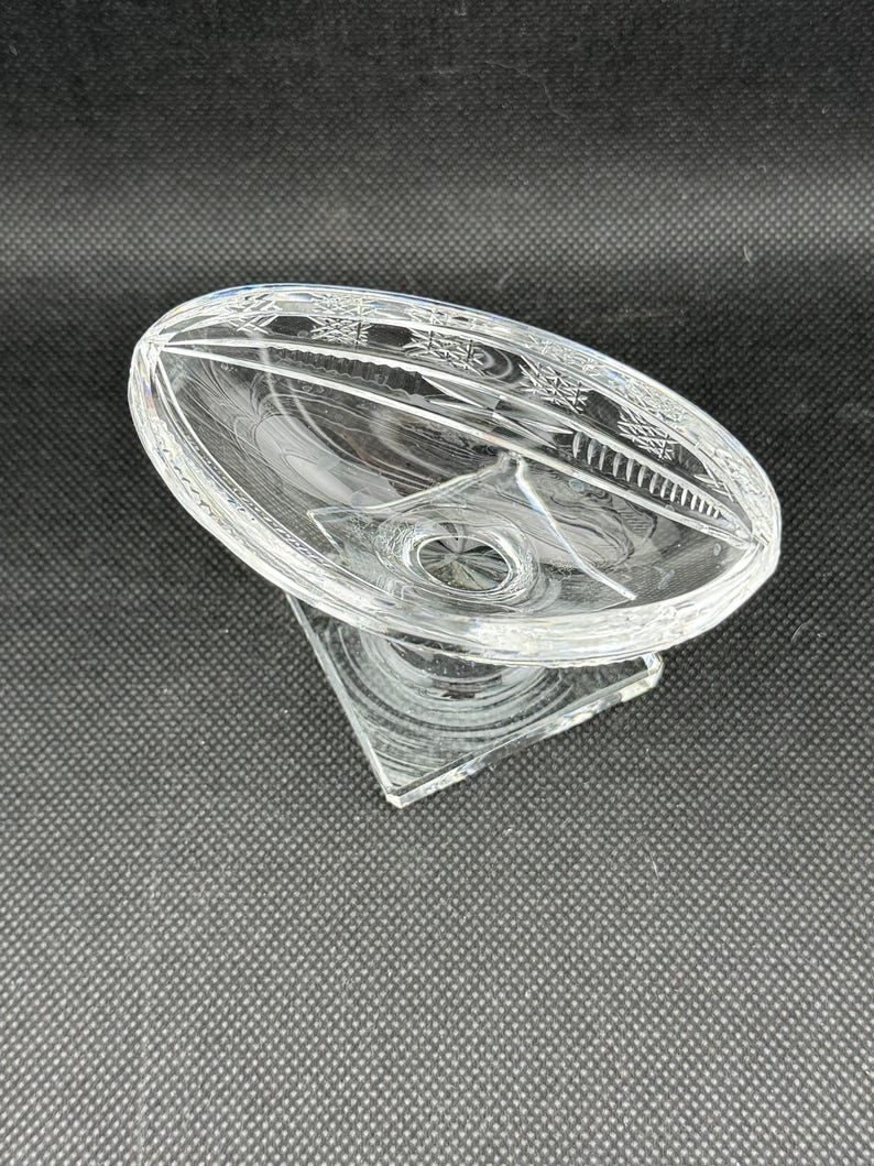 May include: A clear, oval-shaped glass dish with a decorative etched pattern around the rim. The dish sits atop a triangular base, creating a pedestal design. The glass has a reflective quality, catching light.