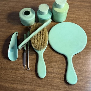 May include: A vintage mint green vanity set, including a hand mirror, brush, comb, jars, and manicure tools. The set is arranged on a wooden surface, showcasing a cohesive color palette and classic design.