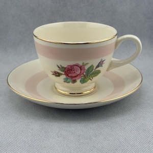 Peut inclure: Un service à thé crème avec une rayure rose et une bordure dorée. La tasse à thé présente un motif floral de rose. La soucoupe a une rayure rose assortie et une bordure dorée. L'ensemble est sur une surface gris clair.