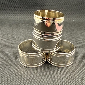 May include: A set of four silver napkin rings. Three rings are arranged in a triangular shape, with one ring stacked on top. The rings have a polished finish with a textured band and a smooth rim. The background is dark.