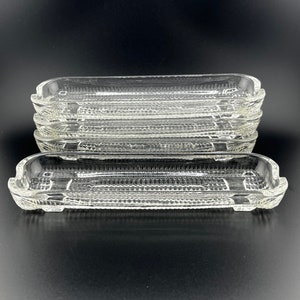 May include: A stack of six clear glass rectangular serving dishes with a textured surface. The dishes are stacked on a black surface.