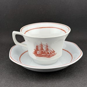 May include: A white teacup and saucer with a red rim and a red ship design. The ship is a three-masted sailing ship with sails.
