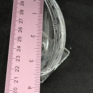 May include: A clear, decorative glass dish with etched floral patterns. The dish is approximately 3 inches (7.6 cm) tall and has a square base. The dish is positioned next to a pink ruler.