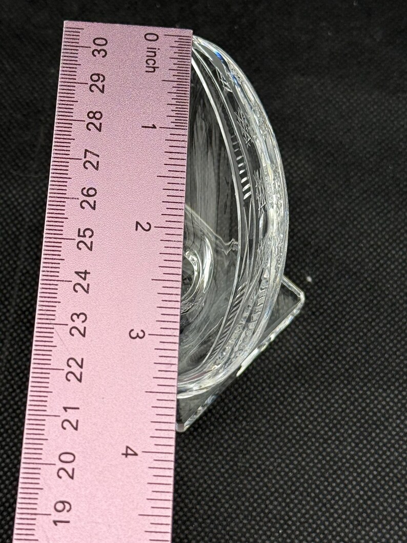 May include: A clear, decorative glass dish with etched floral patterns. The dish is approximately 3 inches (7.6 cm) tall and has a square base. The dish is positioned next to a pink ruler.