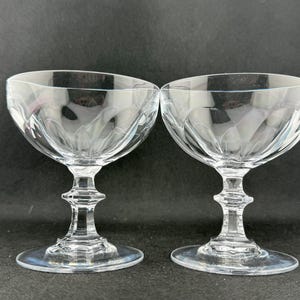 May include: Two clear champagne coupe glasses with etched bowl patterns. The stems have a detailed design, and the bases are round. The glasses are on a dark background. The glasses are ideal for serving champagne.
