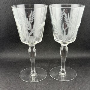 May include: Two clear wine glasses with etched wheat stalk designs. The glasses have a flared bowl, a slender stem, and a round base. The wheat design is etched into the glass, creating a delicate and elegant look. Perfect for serving wine.