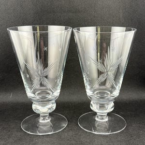 May include: Two clear, stemmed glasses with a starburst design etched into the glass. The glasses have a flared bowl, a rounded stem, and a circular base. The starburst design is repeated on each glass.