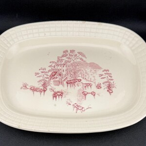May include: Cream-colored, oval serving platter with a red and white landscape design. The platter features a detailed illustration of a building, trees, and a body of water. The rim has a textured, grid-like pattern.