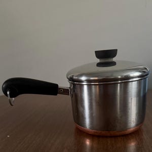 Peut inclure: Une petite casserole en acier inoxydable avec un couvercle et une poignée noire. Le fond de la casserole est cuivré et le bouton du couvercle est noir. La poignée a une boucle. La casserole est sur une surface en bois.