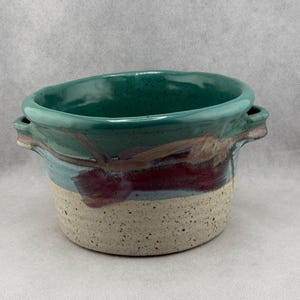 May include: A handcrafted ceramic bowl with a speckled beige base, transitioning to a band of burgundy, light blue, and teal. The top rim is teal, and the bowl has two small handles. This unique piece is perfect for serving or display.