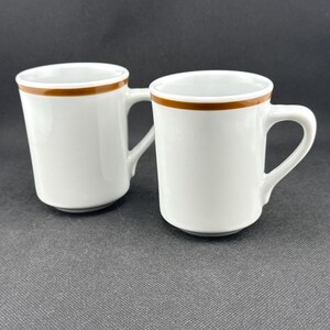 May include: Two white ceramic mugs with a brown band around the top. The mugs have handles.