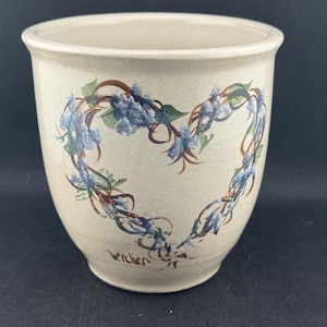 May include: A beige ceramic plant pot featuring a heart-shaped floral design. The heart is made of blue flowers, green leaves, and brown stems. The word "Wendier" is written in cursive on the bottom.