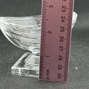 May include: A clear, decorative glass dish with a boat-like shape, resting on a square base. The dish is approximately 3 inches long, with etched details and a textured surface. The ruler shows the measurement in inches.