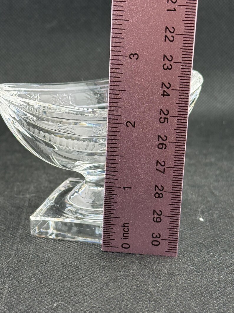 May include: A clear, decorative glass dish with a boat-like shape, resting on a square base. The dish is approximately 3 inches long, with etched details and a textured surface. The ruler shows the measurement in inches.