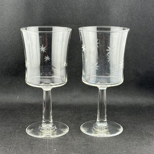 May include: Two clear, stemmed glasses with a starburst pattern etched into the glass. The glasses have a flared top and a round base. The background is a solid black.