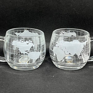 May include: Two clear glass mugs with a frosted design of the world on the front. The mugs have a handle on the right side.