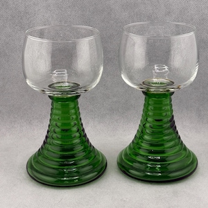 May include: Two clear glass wine goblets with a green, ribbed stem and base. The bowl of the glass is round. The green stem has a flared, conical shape. The glasses are set against a neutral background.