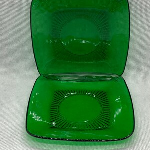 Peut inclure: Deux assiettes carrées en verre vert émeraude. Chaque assiette présente une surface texturée en relief avec un motif ovale central et des lignes rayonnantes. Les assiettes ont une bordure noire et sont empilées l'une sur l'autre.