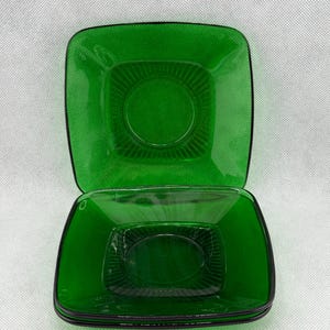 Peut inclure: Une pile de quatre assiettes carrées en verre vert émeraude. Chaque assiette a un centre légèrement surélevé et un motif nervuré. Les bords sont soulignés d'une fine bordure noire. Les assiettes sont empilées sur une surface blanche.
