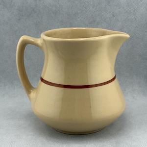 May include: A cream-colored ceramic creamer with a single brown stripe around the middle. The creamer has a rounded body and a small spout.