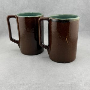 May include: Two brown ceramic mugs with rectangular handles. The interior of each mug is a teal color, with a matching teal rim. The mugs are displayed on a light gray surface.