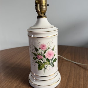 May include: A vintage white ceramic table lamp with a floral rose design. The lamp features a brass neck and a cylindrical base adorned with pink roses and gold trim. The lamp is approximately 12 inches tall.
