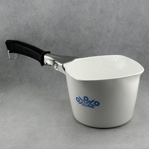 May include: White saucepan with a black handle and a silver metal bracket. The saucepan features a blue floral design. The handle has a metal loop. The saucepan is empty.