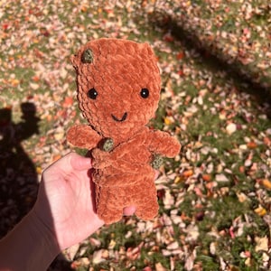 May include: A handmade, plush, brown doll with a smiling face, black eyes, and small green accents. The doll has arms and legs and is made of a soft, textured material. The doll is a cute, handcrafted item.
