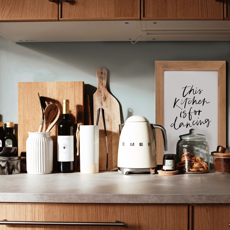 May include: A white electric kettle with a black handle and a silver knob on a wooden countertop. The kettle is in front of a framed sign that reads "This kitchen is for dancing".