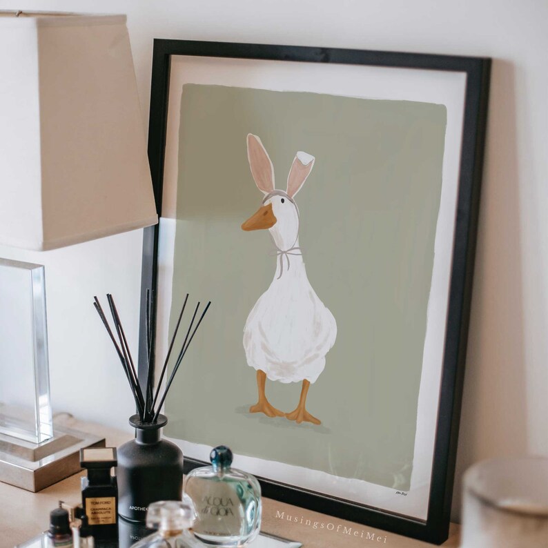 May include: A framed illustration of a white goose wearing bunny ears. The goose is standing on a green background. The frame is black and the illustration is titled "Musing of Mei Mei".