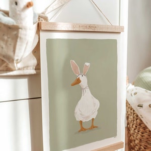 May include: A framed illustration of a white duck wearing pink bunny ears on a green background. The frame is made of wood and has a hanging wire. The text "Little Dutch" is printed on the frame.