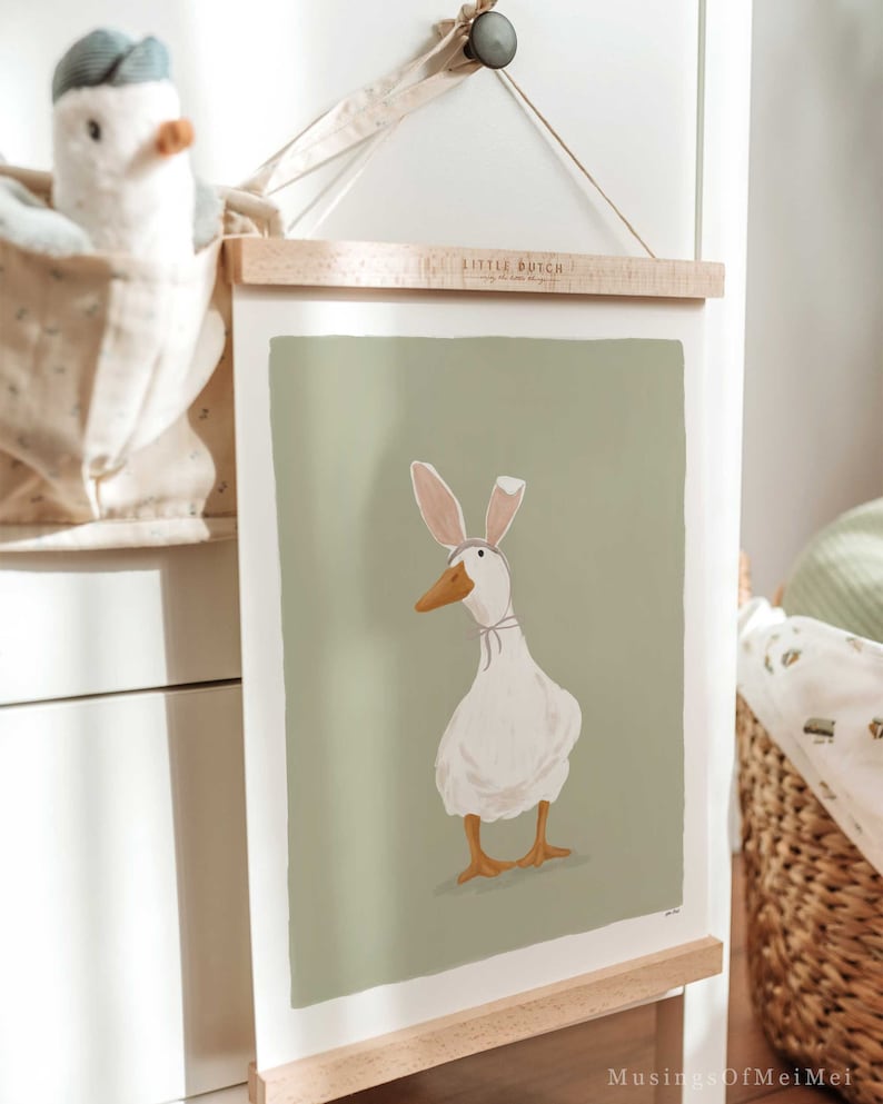 May include: A framed illustration of a white duck wearing pink bunny ears on a green background. The frame is made of wood and has a hanging wire. The text "Little Dutch" is printed on the frame.