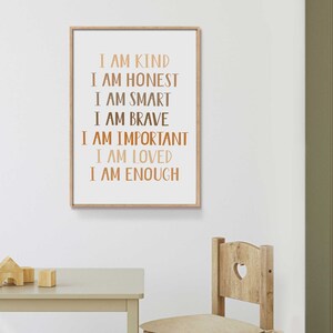 May include: A framed print with the words "I AM KIND, I AM HONEST, I AM SMART, I AM BRAVE, I AM IMPORTANT, I AM LOVED, I AM ENOUGH" in various shades of brown. The print is in a light wood frame and hangs on a white wall.