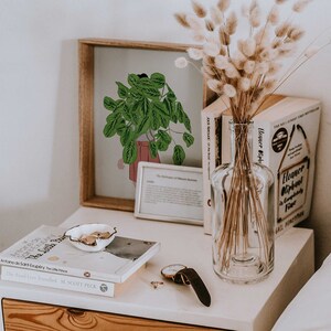 May include: A wooden framed print of a green plant in a pink pot, a clear glass bottle with dried flowers, and a stack of books on a white bedside table. A watch and small dish are also on the table.