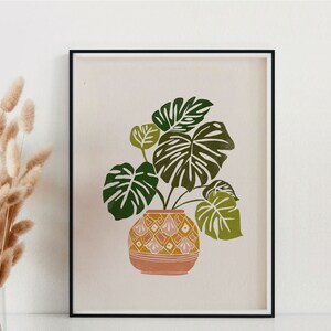 May include: A framed print of a green monstera plant in a pink and yellow patterned pot on a light pink background.