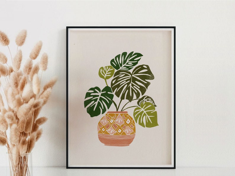 May include: A framed print of a green monstera plant in a pink and yellow patterned pot on a light pink background.