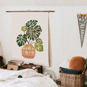 May include: A beige canvas wall tapestry with a green monstera plant in a pink and yellow patterned pot. The tapestry is hanging on a wall with a wooden rod.