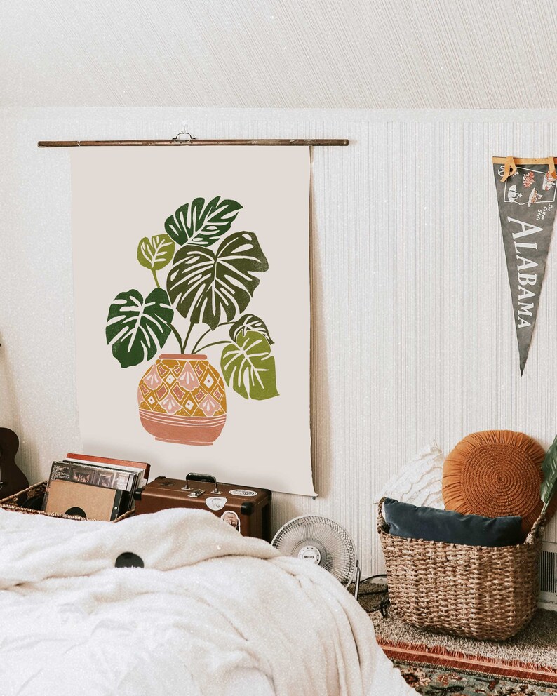 May include: A beige canvas wall tapestry with a green monstera plant in a pink and yellow patterned pot. The tapestry is hanging on a wall with a wooden rod.