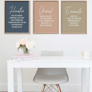 May include: Three motivational posters with a light brown wood frame. The posters have a light blue, light brown, and light gray background. The text on the posters reads "Hustle", "Grind", and "Execute". The posters are hanging on a white wall above a white desk with a light gray chair and a vase of white and pink flowers.